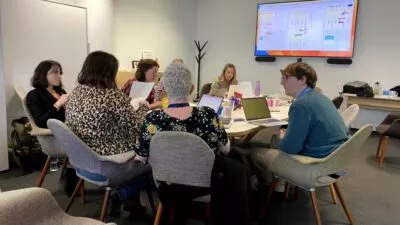A photo of a group of 7 people sat around a table looking at a big screen and talking