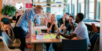 A group of people at UX Brighton creating an 'impact map'. They are sat around a table putting sticky notes on a large piece of paper.