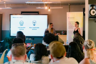 Nicola and Hannah presenting to the audience at UX Brighton. On the board behind them it says 'What is the value? Less harmful services and more sustainable services'