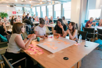A group of people at UX Brighton creating an 'impact map'. They are sat around a table putting sticky notes on a large piece of paper.