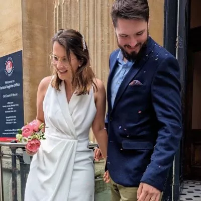 A photo of Amy and Mark, having just got married. Amy is wearing a white dress and carrying a small bunch of peonies. Mark is wearing a blue suit. They're both covered in confetti and smiling