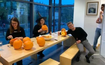 A photo of 3 people carving pumpkins. Charlotte is in the middle, wearing a witches hat and holding up her carved pumpkin