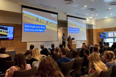 Amanda and Mark stood in front of a podium presenting to a conference hall full of people. Behind them, there are two screens that read 'Mind the gap - how to get more from Discovery and Alpha'.