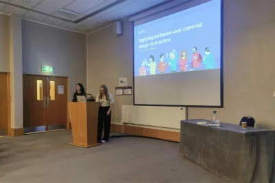 Chloe and Megan stood in front of a podium. Behind them is a screen that says 'Applying inclusive user centred design in practice'