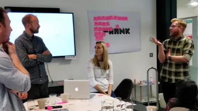 Ollie talking to some of the team working on a Talk to Frank Project. They are stood around a table and in the background there is a board with sticky notes on it.