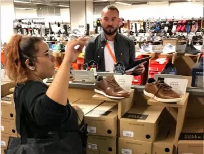 A photo of a man in a shoe shop observing a woman working