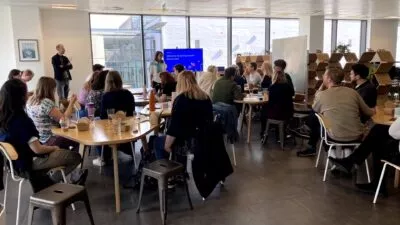 A photo of around 20 employees sat as a group in an office watching someone presenting at the front of the room