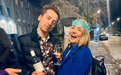A photo of David with his colleague Ashleigh after the cxpartners christmas party. He's holding a bottle of wine and Ashleigh is holding a glass of wine, wearing a paper crown from a Christmas cracker