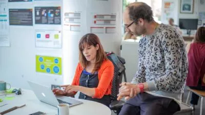 A photo of a woman sat at a desk in an office showing a man something on her laptop screen