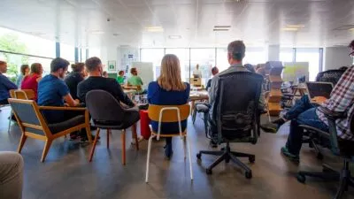 A group of people in an office sat on chairs listening to someone talking at the front