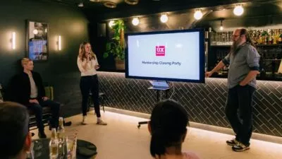 A photo of a woman stood at the front of the room presenting from a big screen.