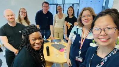 A photo of Bea, Henry, Chloe, James, Natale, Nicola, Sophie and Julie stood around a table in the cxpartners office smiling
