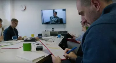 A photo of Stakeholders observing a customer being interviewed by a user researcher. They are in a separate room, watching the interview on a big screen