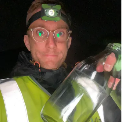 Sam, the author, in a high-viz jacket holding an empty bottle of milk. It's night time and he has a head-torch on.